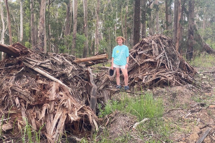 A woman in a bright pale blue t-short stands next to piles of bark in a forest.