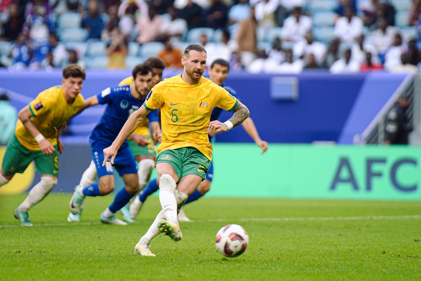 A soccer player wearing yellow and green kicks the ball while his team-mates and opposition wearing all blue run behind him