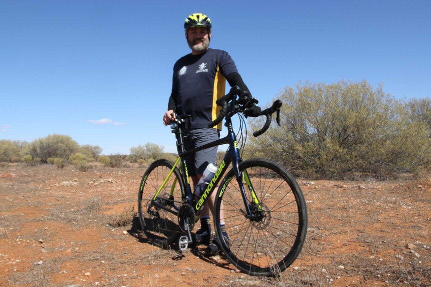 A man stands with his bike in red dirt and bush.