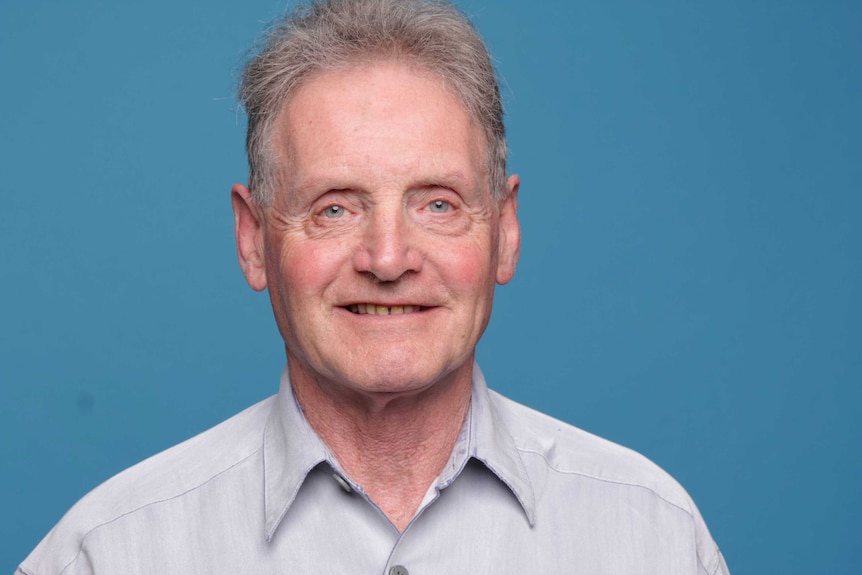 Man in pale suit shirt, before a blue background, smiles slightly.