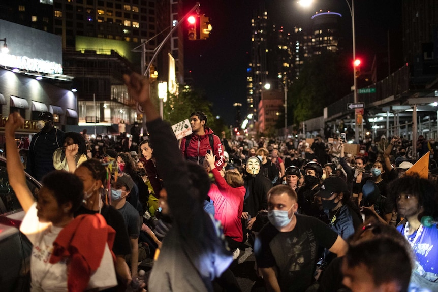 A massive group of protesters, some with fists raised, stand on a long street in the night.