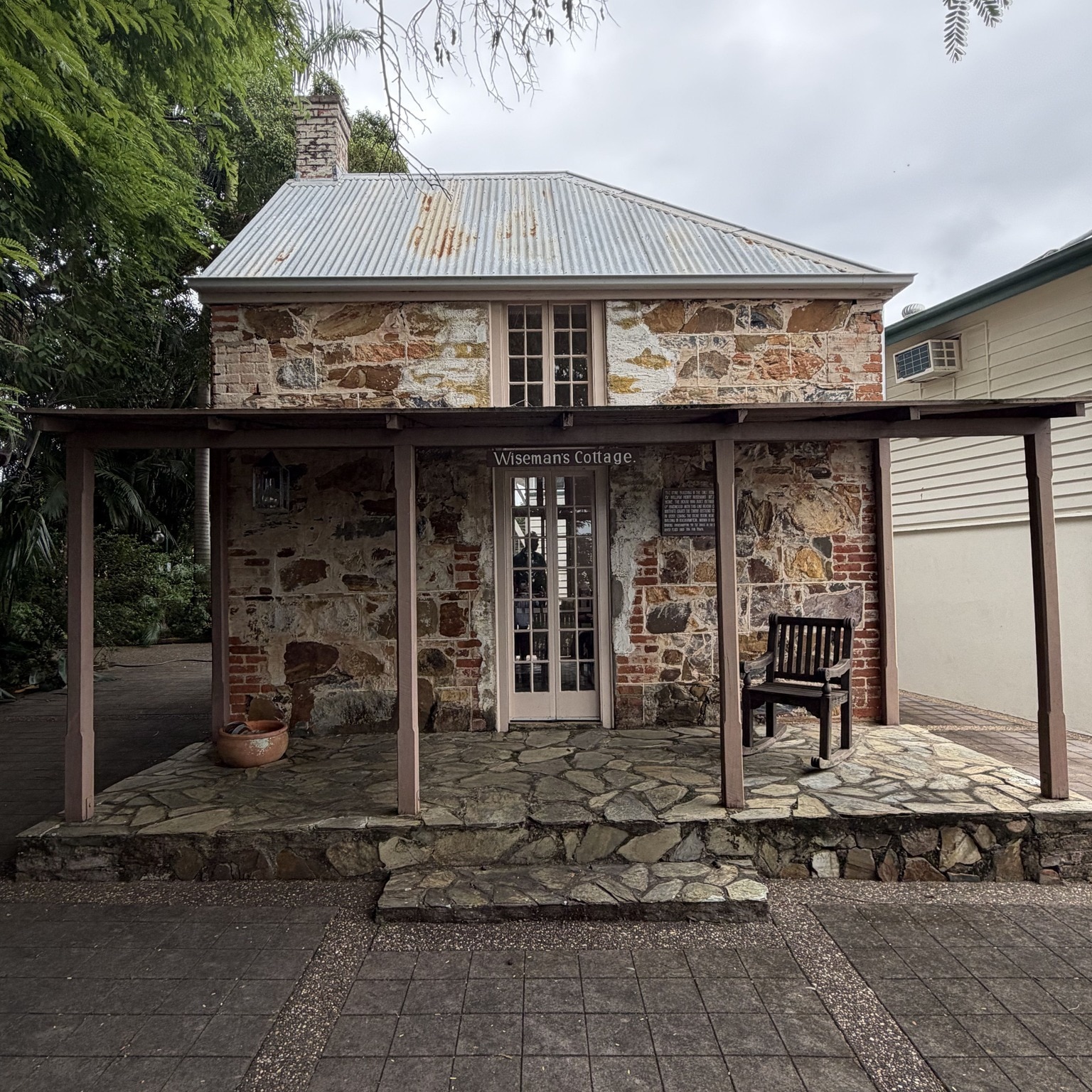 Stone brick cottage structure in garden with tall roof and verandah with wooden chair