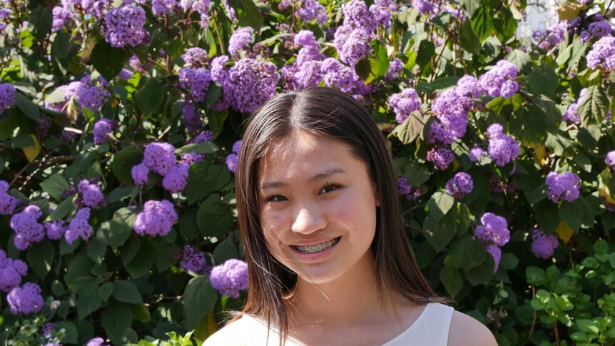 A young woman wearing a white top, stands in front of purple flowers, smiling.