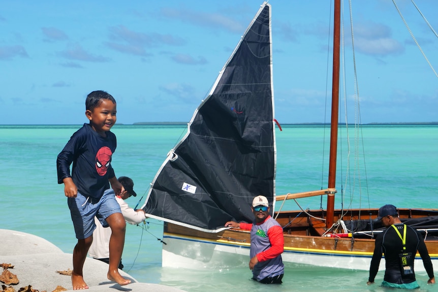 A young boy wearing a spider man shirt with a big grin runs, a sail boat in the background on blue water.
