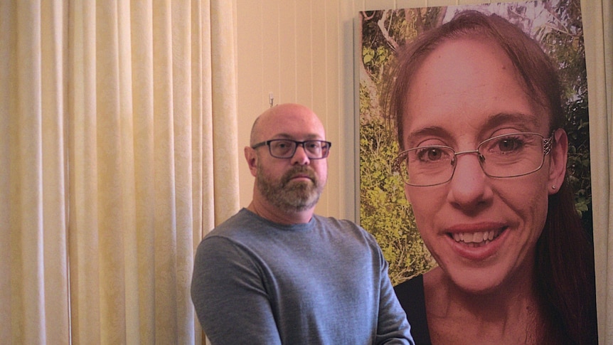 A man with glasses stands near a large photo of a smiling woman that hangs on a wall in a home with white walls.