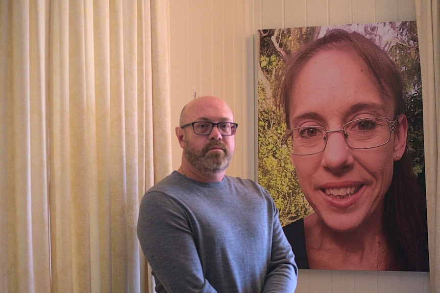 A man with glasses stands near a large photo of a smiling woman that hangs on a wall in a home with white walls.