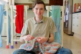 Jorden looks to the camera as he crochets a red, blue and white blanket while sat in his kitchen with projects behind.