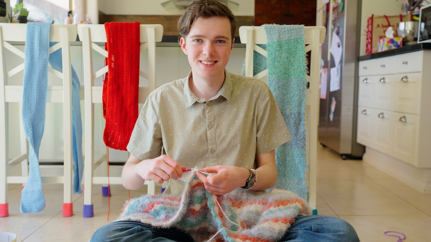 Jorden looks to the camera as he crochets a red, blue and white blanket while sat in his kitchen with projects behind.