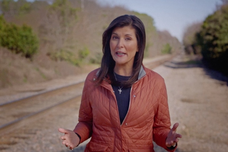 A woman in a brown jacket stands on train tracks with her hands out