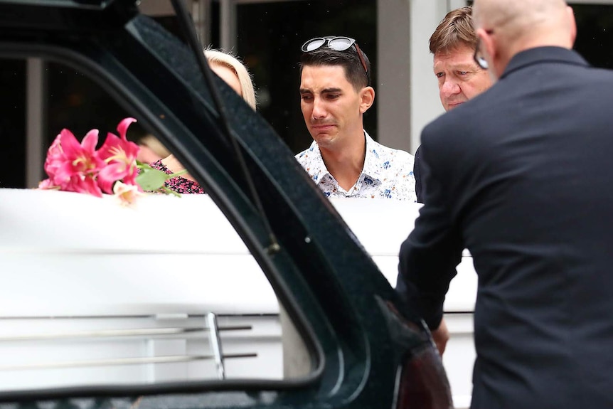 A distraught man watches as a coffin is placed inside a hearse