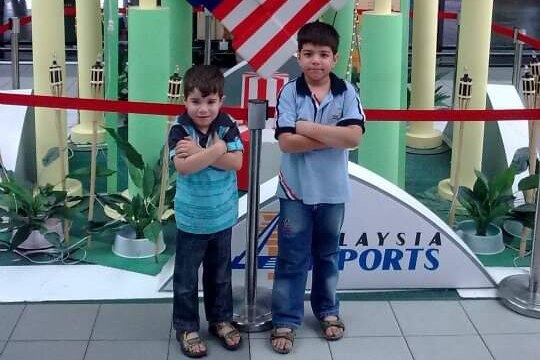 Two young boys stand in front of a Malaysian flag at an airport.