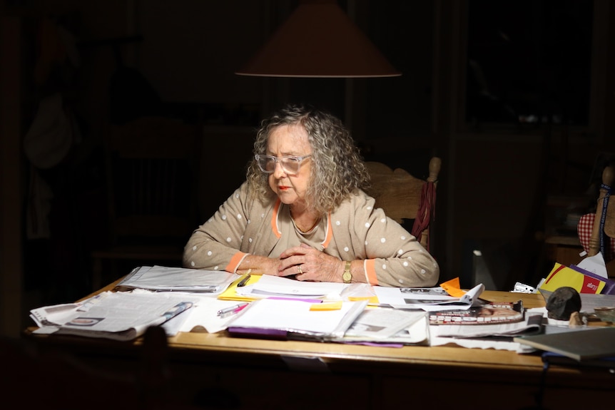 A woman sits at a kitchen table under the light of an overheat light surrounded by documents and paperwork