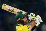 A male cricketer in a green and gold shirt, green helmet, in the follow through of a swing