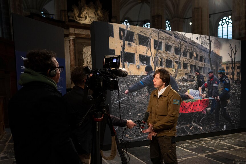 A young man in a brown jacket stands in front of a large printed copy of a war photograph, speaking to reporters holding mics.