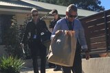 Three officers, including one carrying a large paper bag, walk down steps outside a modern, suburban home.
