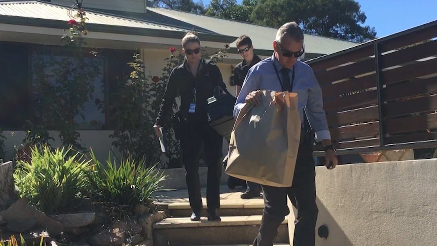 Three officers, including one carrying a large paper bag, walk down steps outside a modern, suburban home.