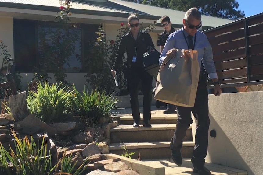 Three officers, including one carrying a large paper bag, walk down steps outside a modern, suburban home.