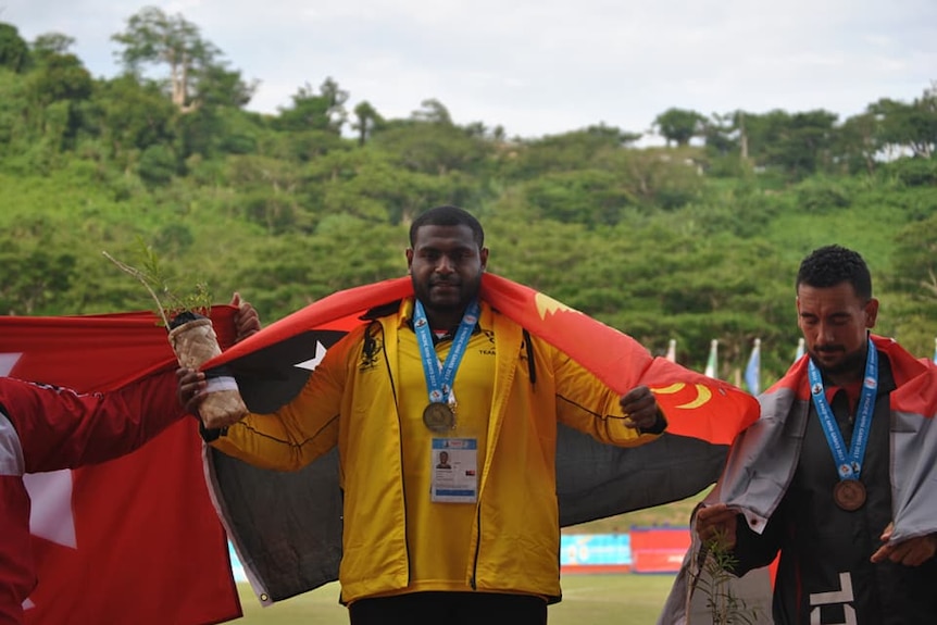 A man stands on a podium wearing a flag with a gold medal around his neck next to another man