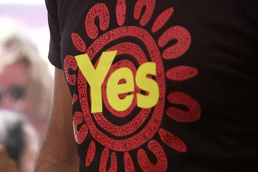 The torso of a person wearing a dark-coloured T-shirt with "Yes" printed on it in bright lettering.