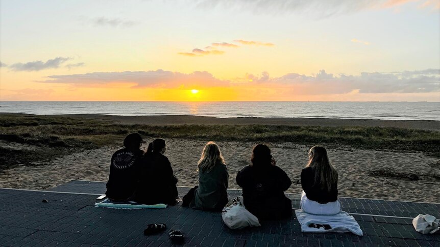 A group of people sit at the beach watching a sunrise, all with backs to the camera.