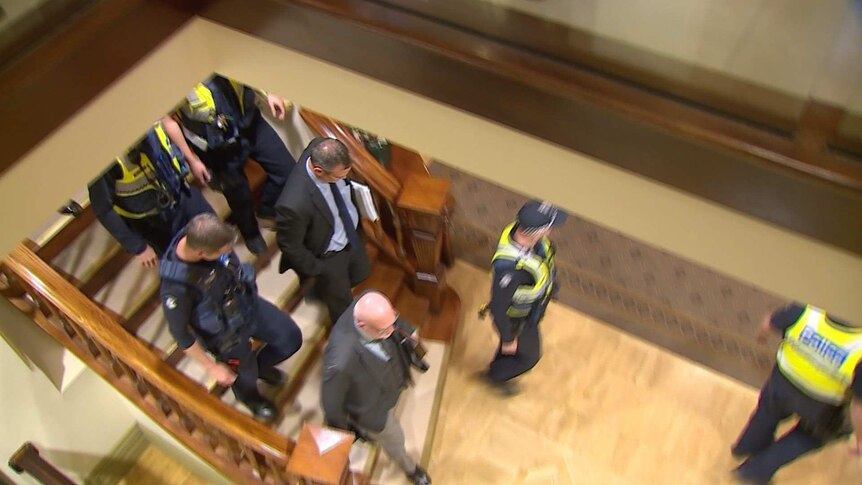 A man is surrounded by police, walking down stairs.