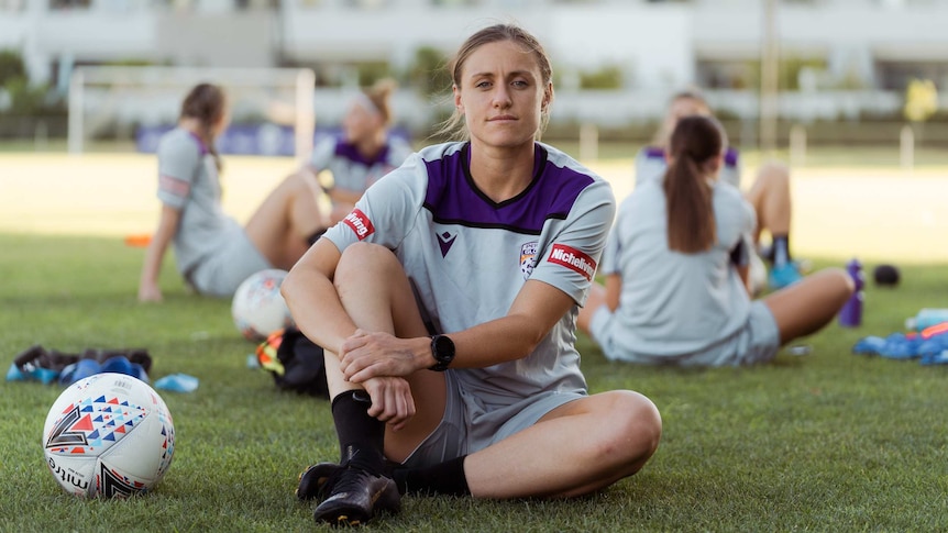 A woman wearing a sports jersey sits on a sports field with a soccer ball next to her