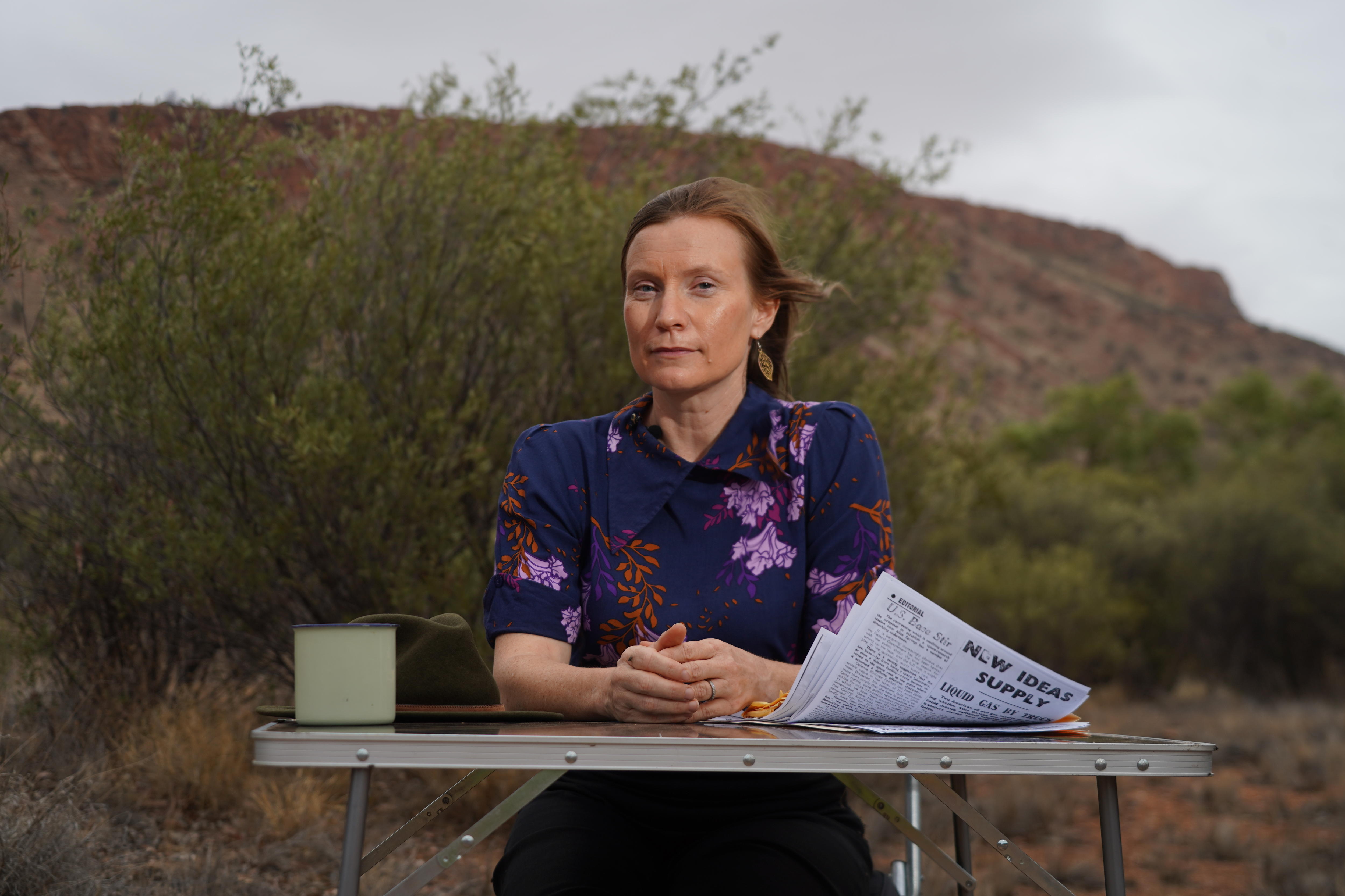 A woman sits at an outdoor table in the outback with sheets of papers in front of her and a hill in the background.. 