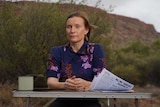 A woman sits at an outdoor table in the outback with sheets of papers in front of her and a hill in the background.. 