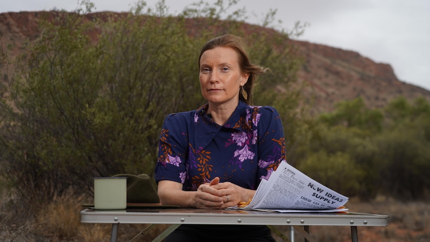 A woman sits at an outdoor table in the outback with sheets of papers in front of her and a hill in the background.. 