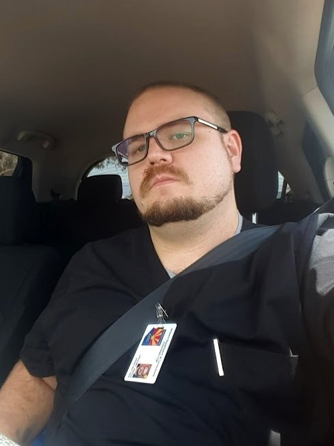 Josh Gregory leans back in a car seat in a selfie photo, wearing black scrubs and a nursing badge.