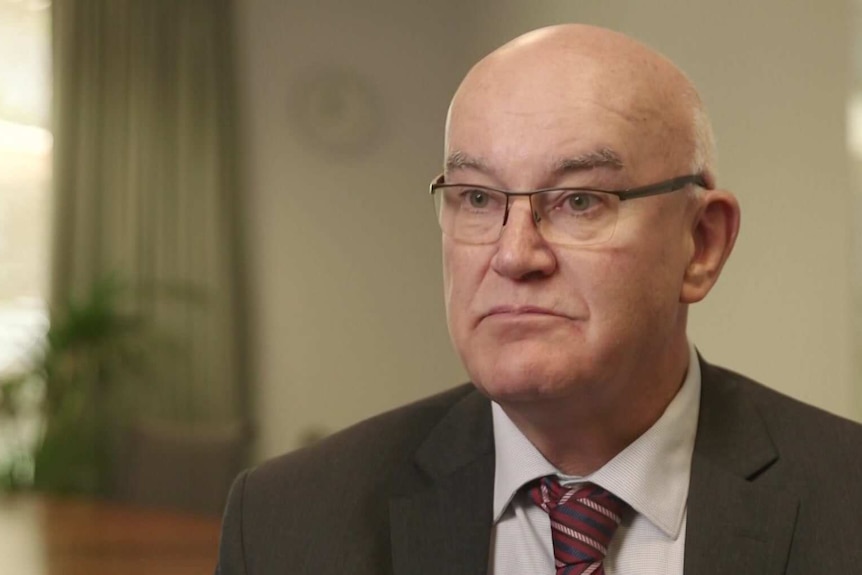 John Skerritt in his office wearing glasses, a black suit and a maroon tie