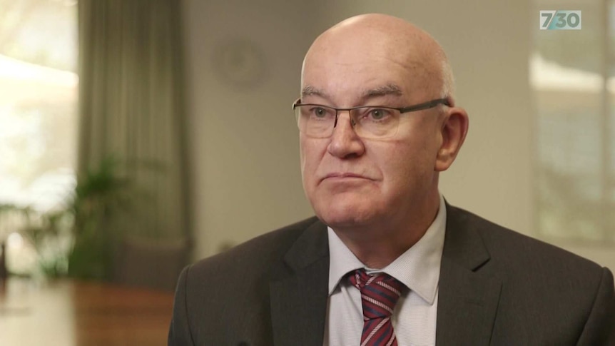 John Skerritt in his office wearing glasses, a black suit and a maroon tie