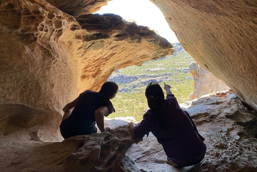 Two children in a cave pointing through a crack with an aerial view of the bush.