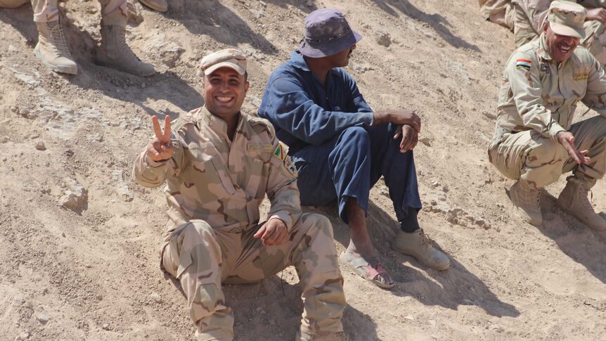 A soldier throws a peace sign while sitting in the sand.