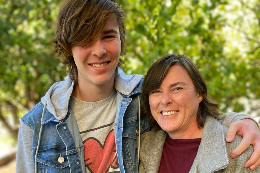 A tall teenager puts his arm round his shorter mum, both of them smiling.