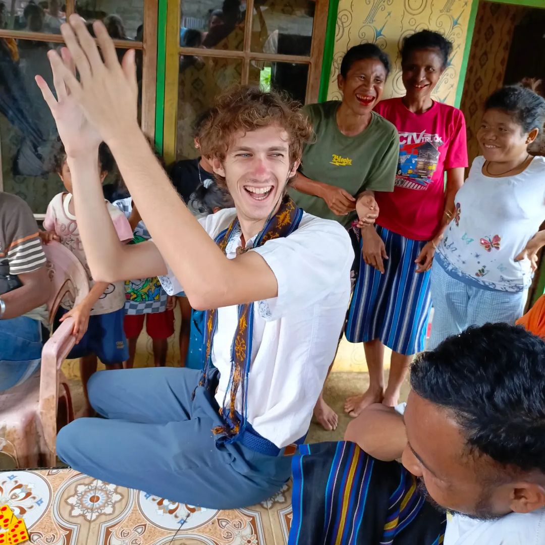 A Caucasian young man smiling and clapping surrounded by his hosts in Timor Leste 