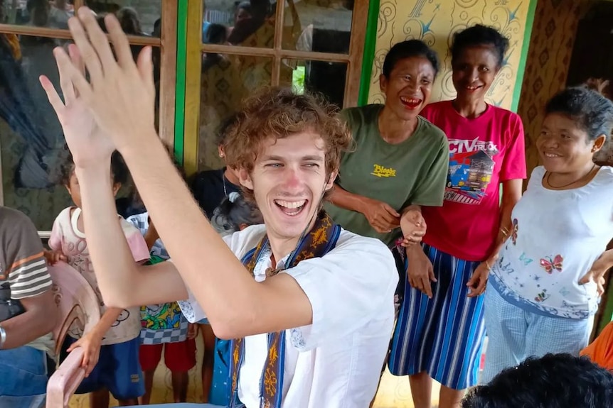 A Caucasian young man smiling and clapping surrounded by his hosts in Timor Leste 