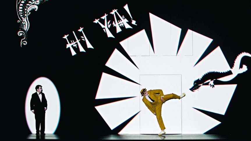 A yellow-suited man kicks at a dragon image on a screen as another suited man looks on.