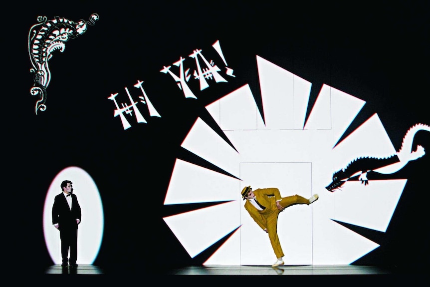 A yellow-suited man kicks at a dragon image on a screen as another suited man looks on.