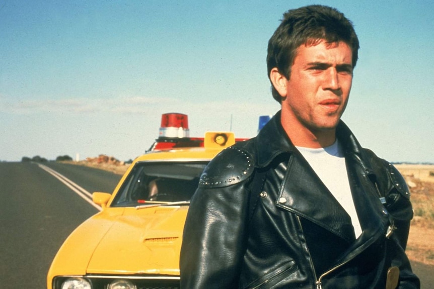 A leather-clad Mel Gibson stands on a desolate, outback road.