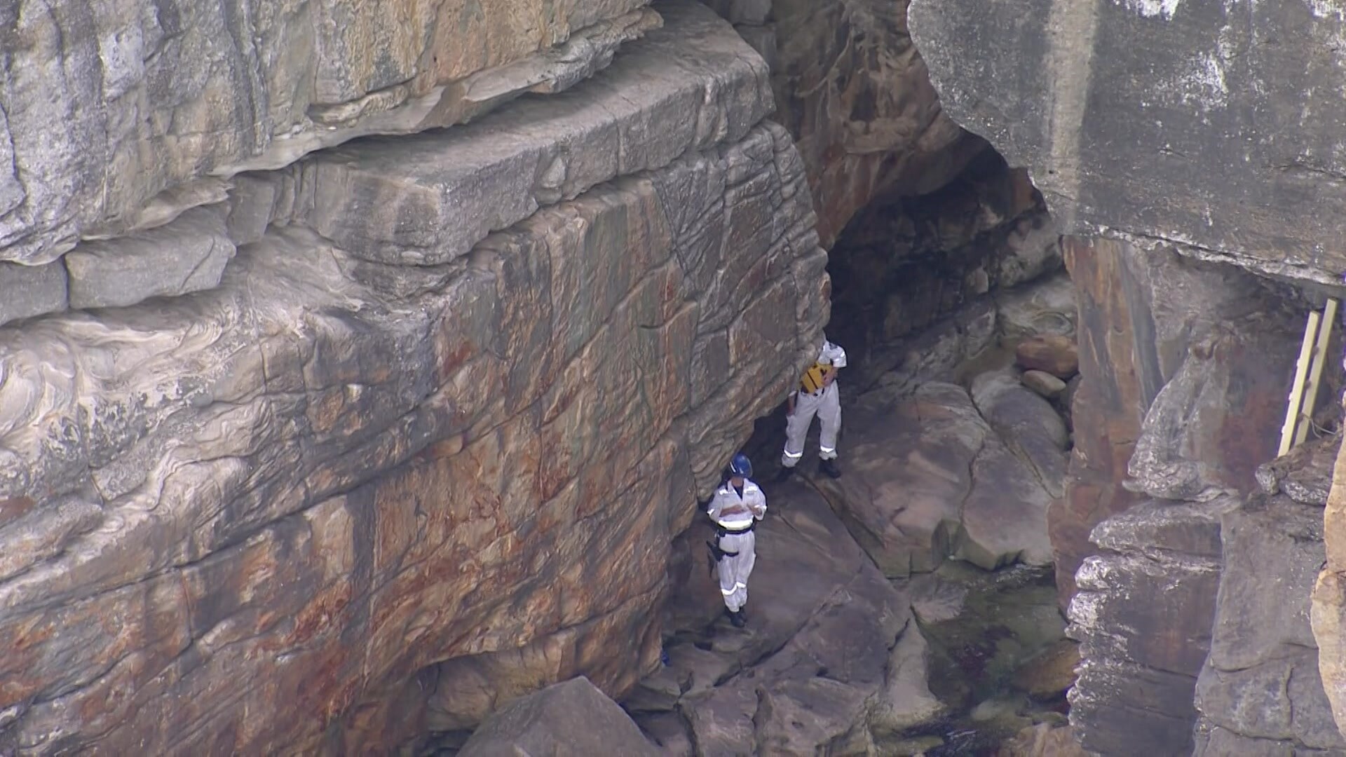 Police rescue officers in white jumpsuits standing on large rocks at the base of steep cliffs at Vaucluse