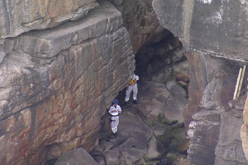 Police rescue officers in white jumpsuits standing on large rocks at the base of steep cliffs at Vaucluse