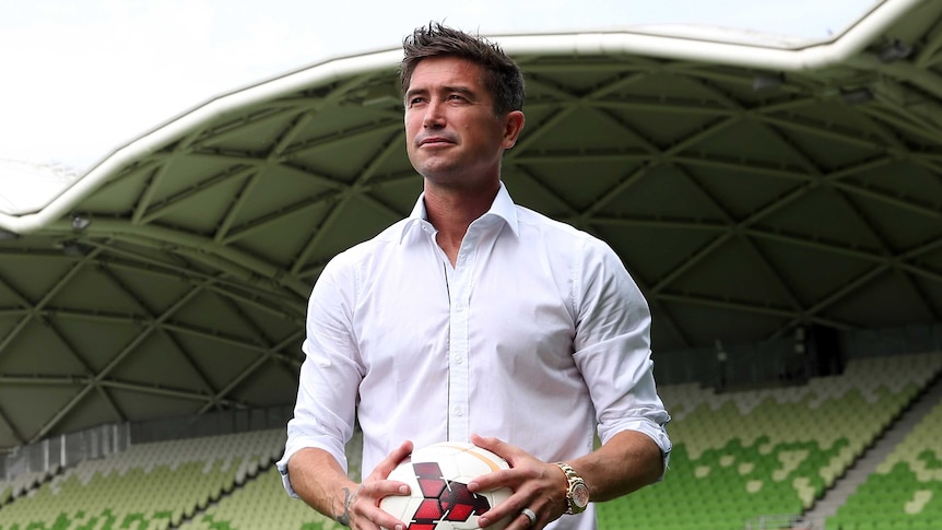 A olive-skinned man with messy dark hair, wearing an open-necked business shirt with rolled-up sleeves stands on soccer pitch