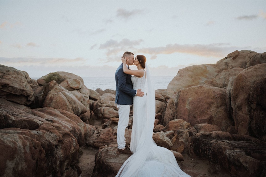 Bride in white dress kisses groom at scenic rocks. 