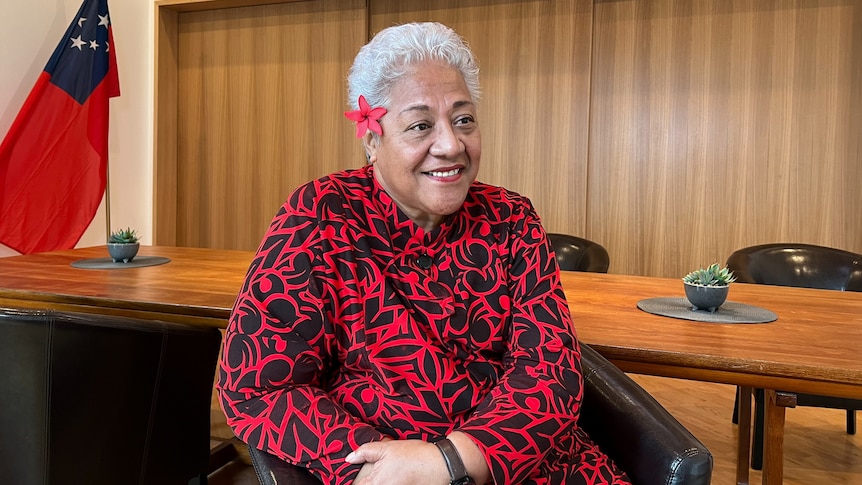 A Samoan woman sitting in red and black patterned outfit with short white hair and red flower behind her ear.