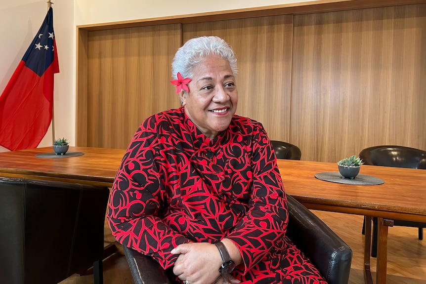 A Samoan woman sitting in red and black patterned outfit with short white hair and red flower behind her ear.