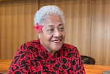A Samoan woman sitting in red and black patterned outfit with short white hair and red flower behind her ear.