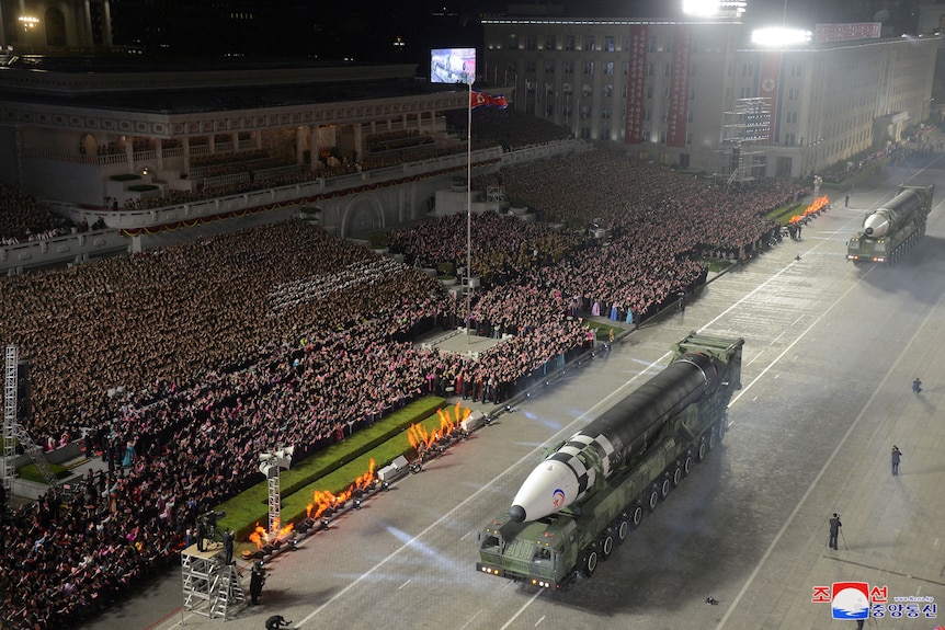 A large missile is displayed on a tank going past a large crowd at night.