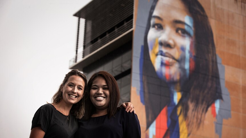 Two women stand together underneath a large mural of one of their faces on the side of a building.
