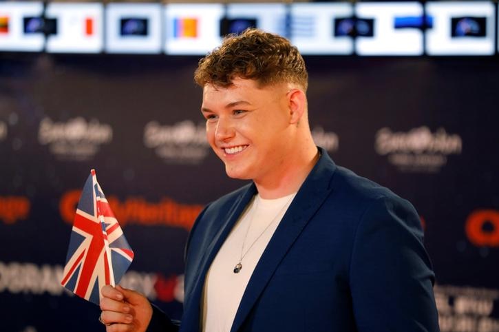 Michael Rice smiles as he holds the UK flag. He is wearing a blue blazer and a white t-shirt.